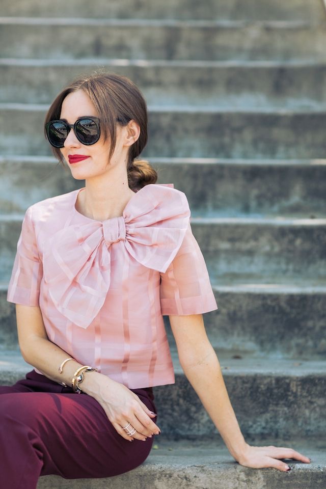 A white lady sitting down wearing a blush pink bow top and burgundy pants