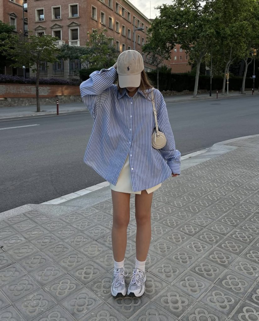 A white lady styling a blue striped baggy shirt with a white short skirt and snaeakers