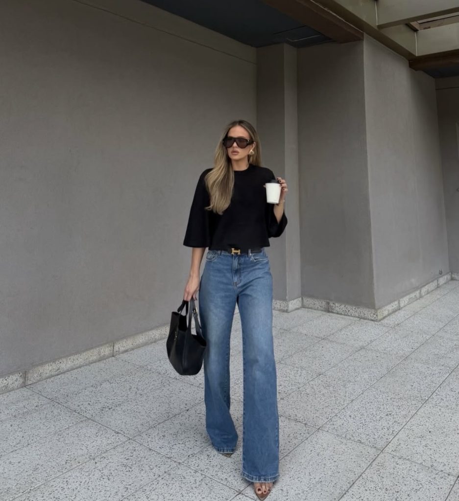 A beautiful woman dressed in casual baggy blue jeans and black tshirt with coffee in one hand and a black handbag in the other.