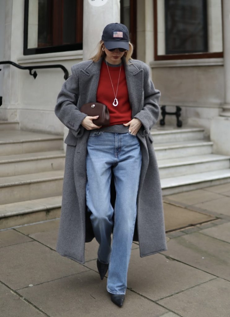 A beautiful woman dressed in casual baggy blue jeans and red tshirt with a big grey trench coat and a face cap.